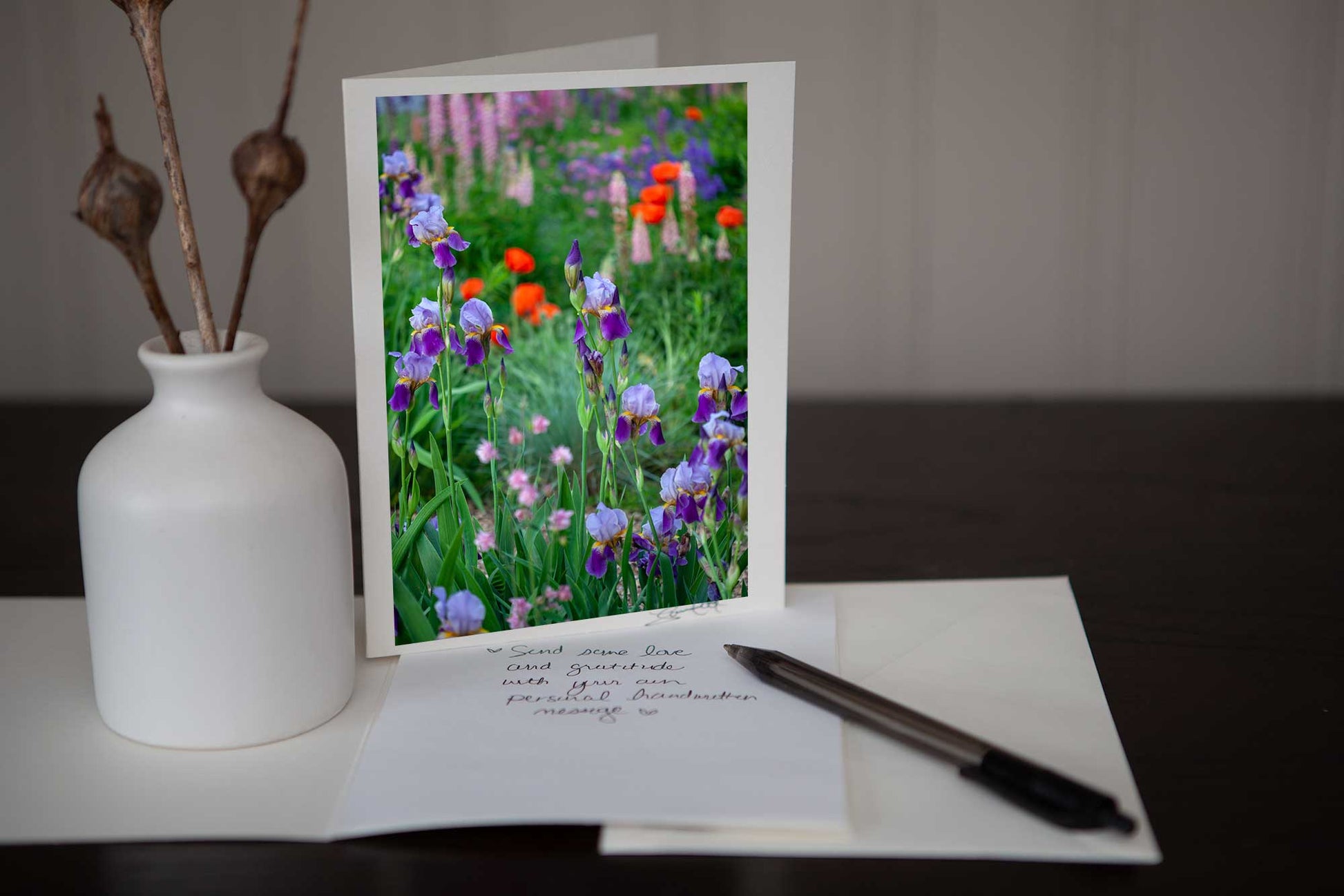 Photo greeting card featuring an image of a close up of purple irises in the garden with some blurred pink lupines in the background along with some red poppies . Photo is mounted to white card stock and signed by the artist Laura Cook in the bottom right corner. 